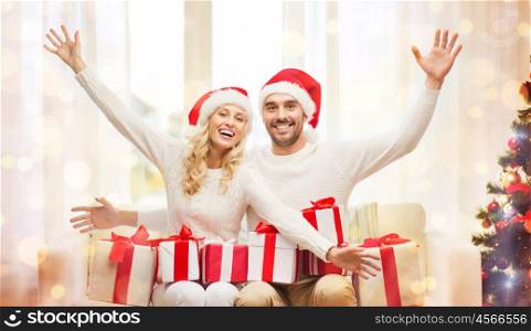 christmas, holidays and people concept - happy couple in santa hats with gift boxes sitting on sofa at home over lights