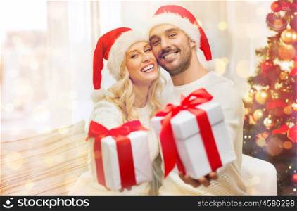 christmas, holidays and people concept - happy couple in santa hats with gift boxes sitting on sofa at home over lights