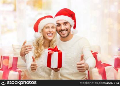 christmas, holidays and people concept - happy couple in santa hats with gift boxes sitting on sofa and showing thumbs up at home over lights