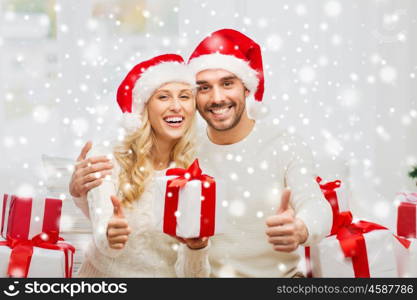 christmas, holidays and people concept - happy couple in santa hats with gift boxes sitting on sofa and showing thumbs up at home