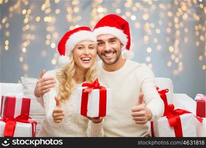 christmas, holidays and people concept - happy couple in santa hats with gift boxes sitting on sofa and showing thumbs up over lights background