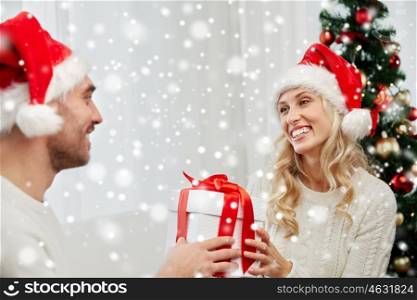 christmas, holidays and people concept - happy couple in santa hats with gift box sitting on sofa at home