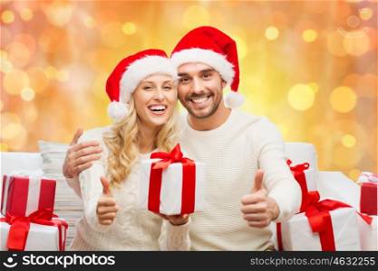 christmas, holidays and people concept - happy couple in santa hats with gift boxes sitting on sofa and showing thumbs up over holidays lights background