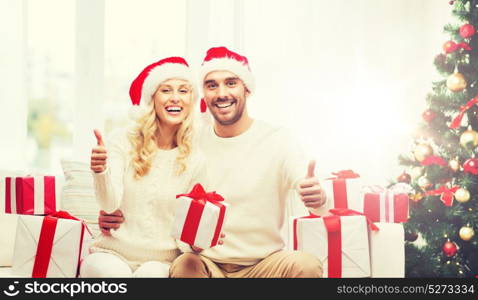 christmas, holidays and people concept - happy couple in santa hats with gift boxes sitting on sofa and showing thumbs up at home. happy couple with christmas gifts and thumbs up