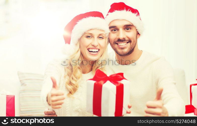 christmas, holidays and people concept - happy couple in santa hats with gift boxes sitting on sofa and showing thumbs up at home. happy couple with christmas gifts and thumbs up