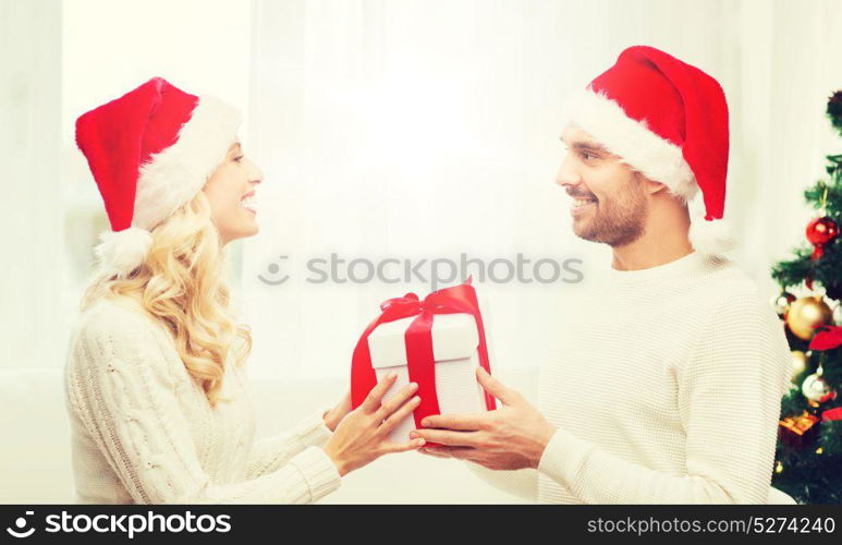 christmas, holidays and people concept - happy couple in santa hats with gift box sitting on sofa at home. happy couple at home with christmas gift box