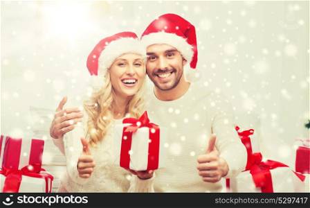 christmas, holidays and people concept - happy couple in santa hats with gift boxes sitting on sofa and showing thumbs up at home. happy couple with christmas gifts and thumbs up