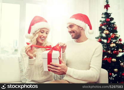 christmas, holidays and people concept - happy couple in santa hats with gift box sitting on sofa at home. happy couple at home with christmas gift box