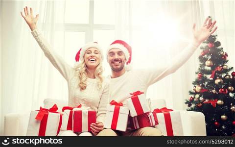 christmas, holidays and people concept - happy couple in santa hats with gift boxes sitting on sofa at home. happy couple at home with christmas gift boxes