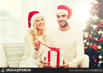 christmas, holidays and people concept - happy couple in santa hats with gift box sitting on sofa at home. happy couple at home with christmas gift box