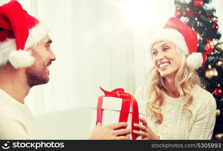 christmas, holidays and people concept - happy couple in santa hats with gift box sitting on sofa at home. happy couple at home with christmas gift box