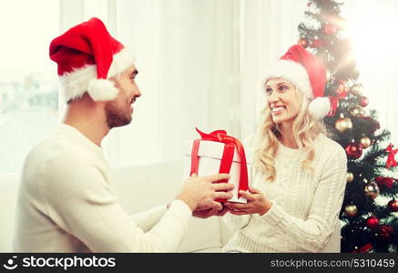 christmas, holidays and people concept - happy couple in santa hats with gift box sitting on sofa at home. happy couple at home with christmas gift box