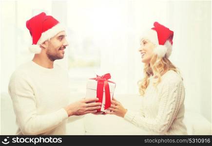 christmas, holidays and people concept - happy couple in santa hats with gift box sitting on sofa at home. happy couple at home with christmas gift box