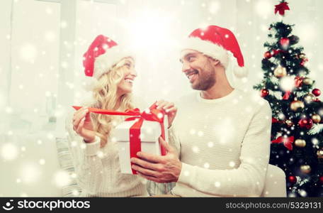 christmas, holidays and people concept - happy couple in santa hats with gift box sitting on sofa at home. happy couple at home with christmas gift box