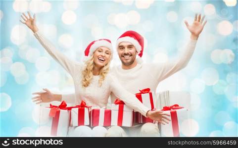 christmas, holidays and people concept - happy couple in santa hats with gift boxes sitting on sofa at home over blue lights background