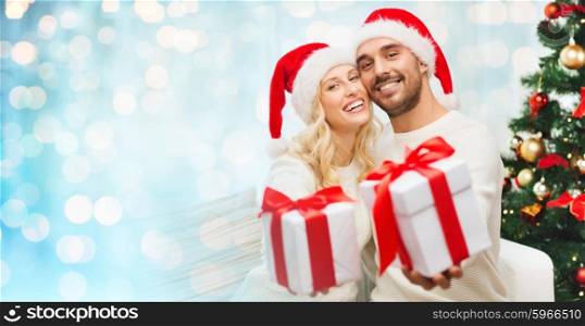 christmas, holidays and people concept - happy couple in santa hats with gift boxes sitting on sofa at home over blue lights background