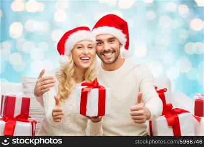 christmas, holidays and people concept - happy couple in santa hats with gift boxes sitting on sofa and showing thumbs up at home over blue lights background