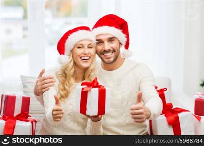 christmas, holidays and people concept - happy couple in santa hats with gift boxes sitting on sofa and showing thumbs up at home