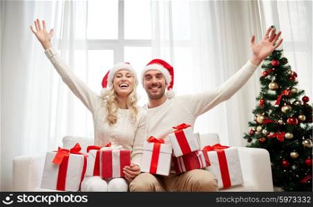 christmas, holidays and people concept - happy couple in santa hats with gift boxes sitting on sofa at home