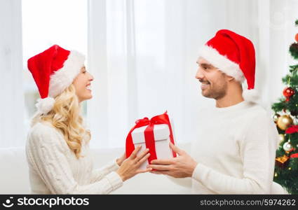 christmas, holidays and people concept - happy couple in santa hats with gift box sitting on sofa at home