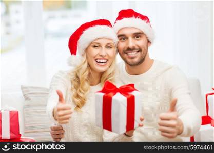 christmas, holidays and people concept - happy couple in santa hats with gift boxes sitting on sofa and showing thumbs up at home