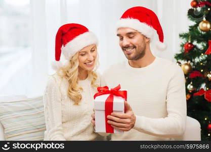 christmas, holidays and people concept - happy couple in santa hats with gift box sitting on sofa at home