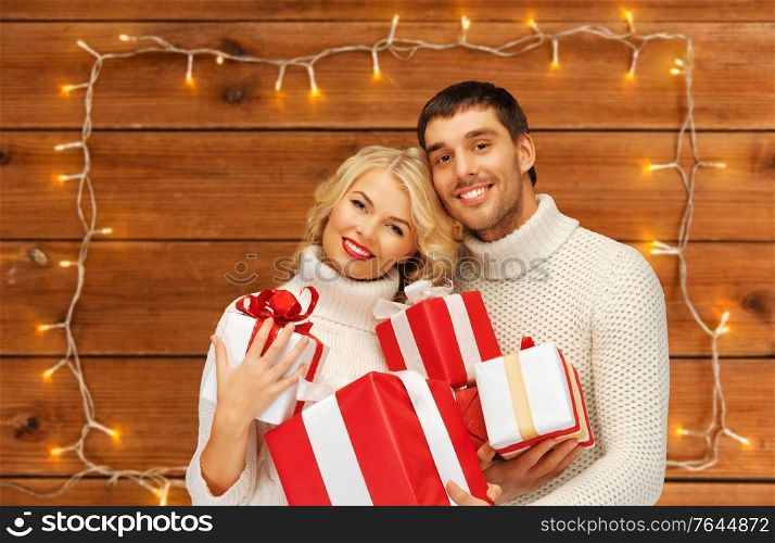 christmas, holidays and people concept - happy couple in sweaters with gift boxes over garland lights on wooden background. happy couple in sweaters with christmas gifts