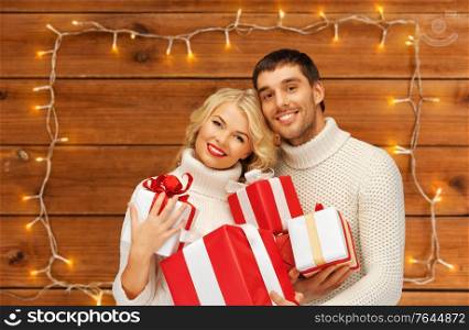 christmas, holidays and people concept - happy couple in sweaters with gift boxes over garland lights on wooden background. happy couple in sweaters with christmas gifts