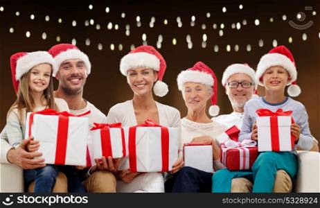 christmas, holidays and people concept - happy family in santa helper hats with gift boxes sitting on couch over garland lights background. happy family in santa hats with christmas gifts