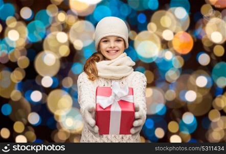 christmas, holidays and people concept - happy girl in winter clothes with gift box over lights background. happy girl in winter clothes with gift box