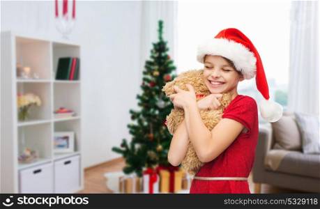 christmas, holidays and people concept - happy smiling girl in santa helper hat with teddy bear at home. happy girl with teddy bear at christmas