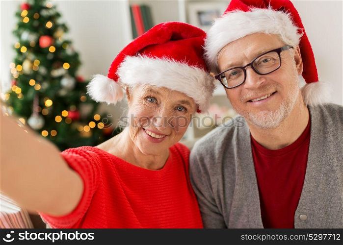 christmas, holidays and people concept - happy smiling senior couple in santa hats taking selfie at home. happy senior couple taking christmas selfie