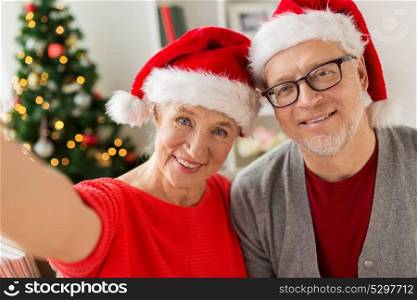 christmas, holidays and people concept - happy smiling senior couple in santa hats taking selfie at home. happy senior couple taking christmas selfie