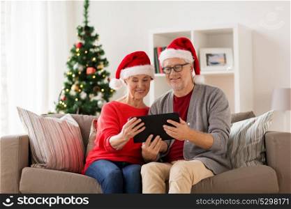 christmas, holidays and people concept - happy smiling senior couple in santa hats with tablet pc computer at home. happy senior couple with tablet pc at christmas