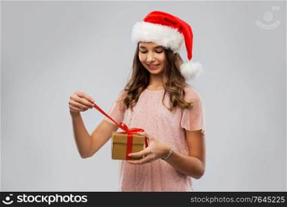 christmas, holidays and people concept - happy smiling teenage girl in santa helper hat opening gift box over grey background. teenage girl in santa hat opening christmas gift