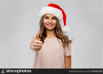 christmas, holidays and people concept - happy smiling teenage girl in santa helper hat showing thumbs up over grey background. happy teenage girl in santa hat showing thumbs up