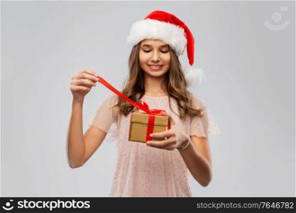 christmas, holidays and people concept - happy smiling teenage girl in santa helper hat opening gift box over grey background. teenage girl in santa hat opening christmas gift