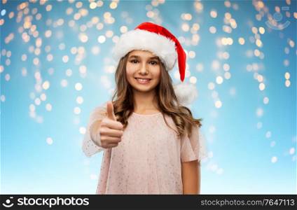 christmas, holidays and people concept - happy smiling teenage girl in santa helper hat showing thumbs up over festive lights on blue background. happy teenage girl in santa hat showing thumbs up
