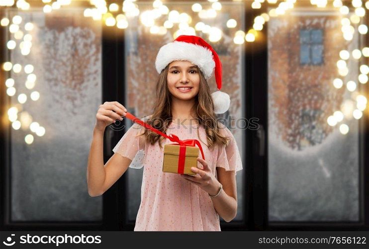 christmas, holidays and people concept - happy smiling teenage girl in santa helper hat opening gift box over garland lights on snowy window background. teenage girl in santa hat opening christmas gift