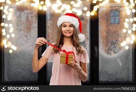 christmas, holidays and people concept - happy smiling teenage girl in santa helper hat opening gift box over garland lights on snowy window background. teenage girl in santa hat opening christmas gift
