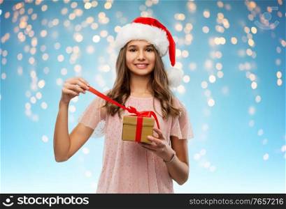 christmas, holidays and people concept - happy smiling teenage girl in santa helper hat opening gift box over lights on blue background. teenage girl in santa hat opening christmas gift