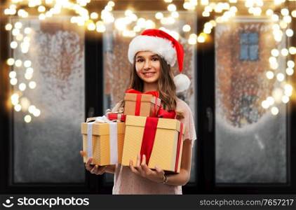 christmas, holidays and people concept - happy smiling teenage girl in santa helper hat holding gift box over garland lights on snowy window background. teenage girl in santa hat with christmas gift