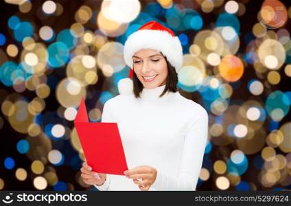 christmas, holidays and people concept - happy smiling woman in santa hat reading greeting card over lights background. happy woman reading christmas greeting card