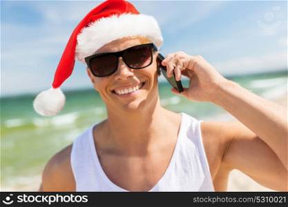 christmas, holidays and people concept - happy smiling young man in santa hat and sunglasses calling on smartphone on beach. man calling on smartphone on beach at christmas