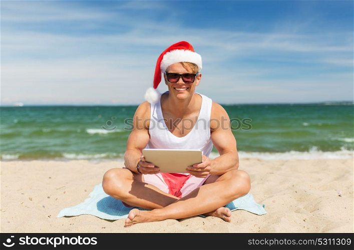 christmas, holidays and people concept - happy smiling young man in santa hat with tablet pc computer sunbathing on beach towel. happy man with tablet pc on beach at christmas