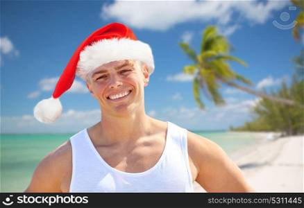 christmas, holidays and people concept - happy smiling young man in santa hat on beach. happy young man in santa hat on beach at christmas