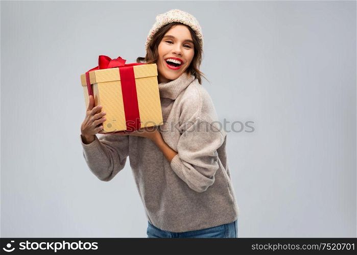 christmas, holidays and people concept - happy smiling young woman in knitted winter hat and sweater holding gift box over grey background. young woman in knitted winter hat holding gift box
