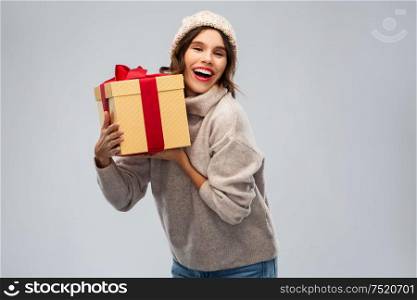 christmas, holidays and people concept - happy smiling young woman in knitted winter hat and sweater holding gift box over grey background. young woman in knitted winter hat holding gift box