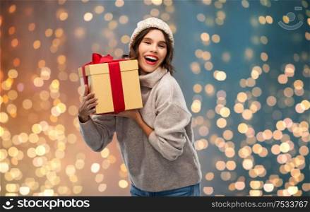christmas, holidays and people concept - happy smiling young woman in knitted winter hat and sweater holding gift box over festive lights background. young woman in knitted winter hat holding gift box