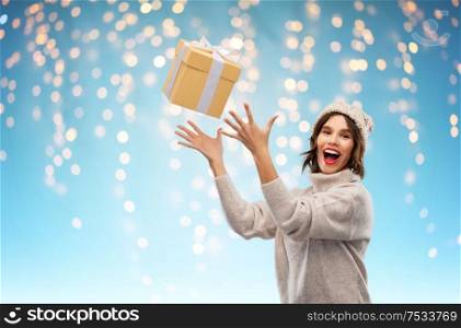 christmas, holidays and people concept - happy smiling young woman in knitted winter hat and sweater catching gift box over festive lights on blue background. young woman in winter hat catching gift box